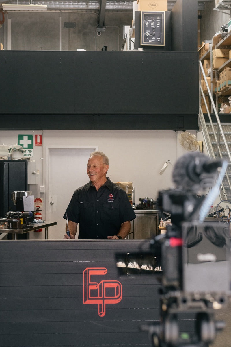 A smiling technician at The Espresso Project service counter, warehouse shelving behind